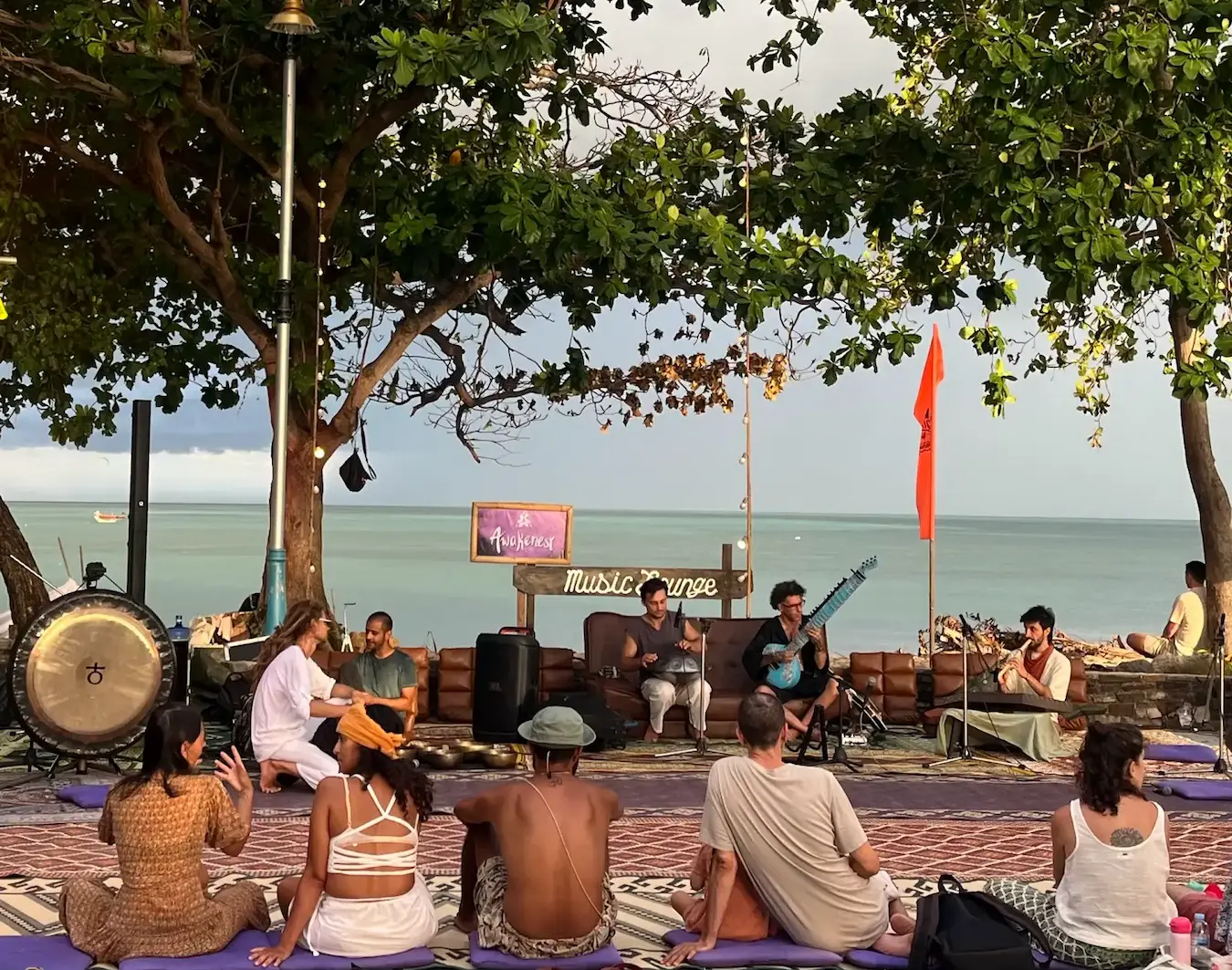 A wide view of Music Lounge by the Sea, a seated gathering on rugs beneath the trees facing the water.