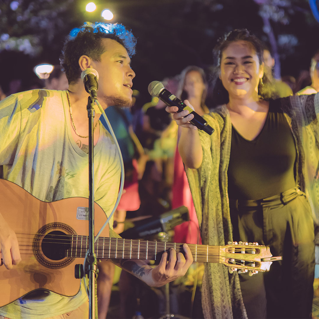 A guitarist and a singer share a microphone under festoon lights at night.